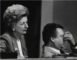 An older white woman wearing a lapel mic and looking tired. A man in the background rubs his eyes. 